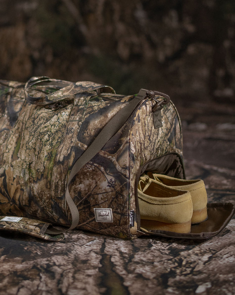 Herschel Supply Co. x Realtree limited-edition collection essential bags, apparel, and accessories arranged together in the APX camouflage pattern.