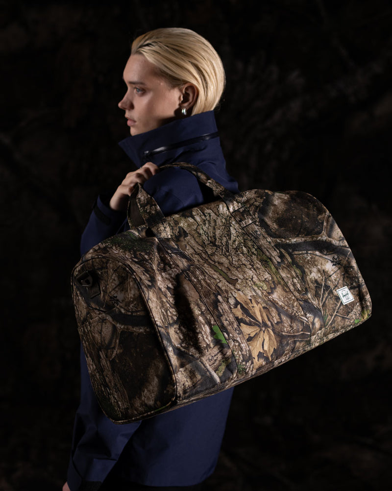 Herschel Supply Co. x Realtree limited-edition collection essential bags, apparel, and accessories arranged together in the APX camouflage pattern.