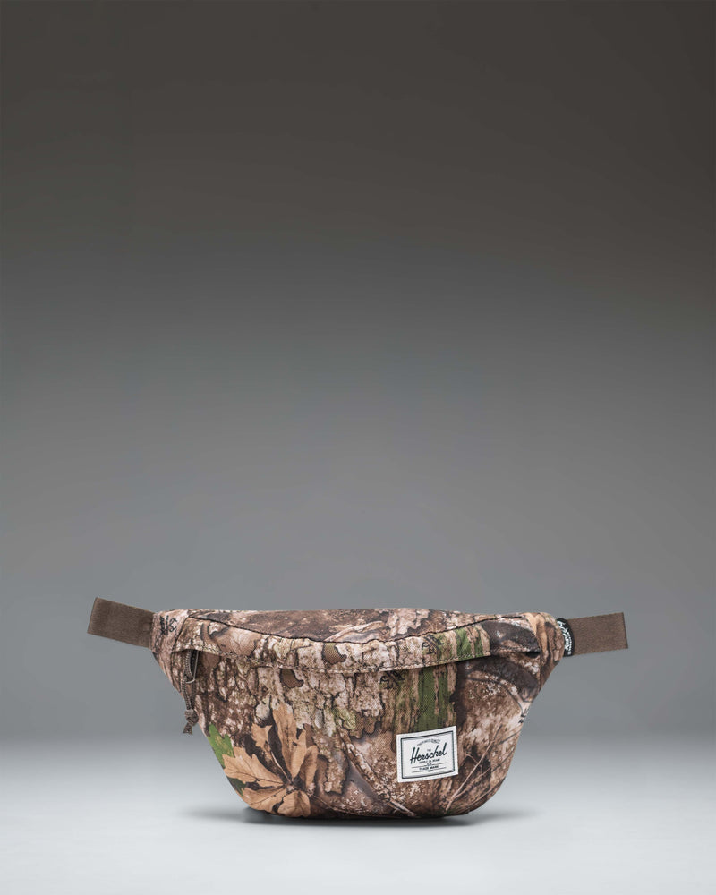 Realtree Herschel Classic Hip Pack in Realtree APX Camo, showing the front zippered pocket and adjustable strap.