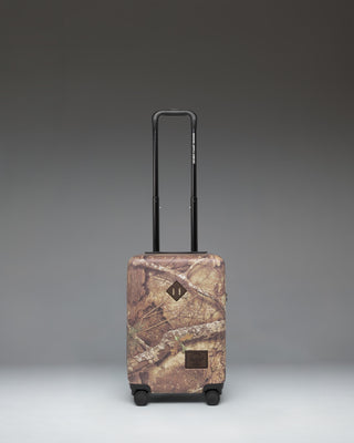 Herschel Heritage Carry On Luggage in Realtree APX Camo pattern, showing the front view with a black zipper and signature logo label.