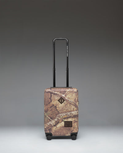 Herschel Heritage Carry On Luggage in Realtree APX Camo pattern, showing the front view with a black zipper and signature logo label.