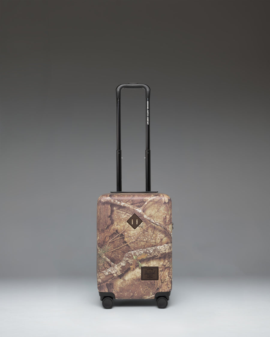 Herschel Heritage Carry On Luggage in Realtree APX Camo pattern, showing the front view with a black zipper and signature logo label.