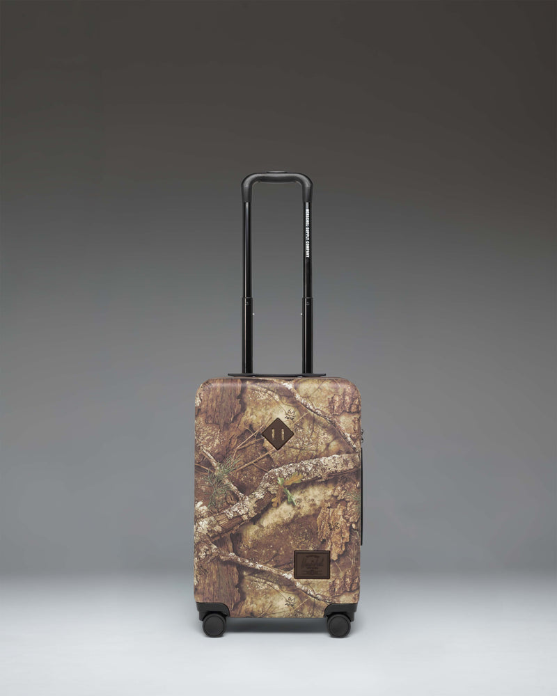 Herschel Heritage Large Carry-On Luggage in Realtree APX Camo pattern, showing the front view of the hard-shell suitcase.
