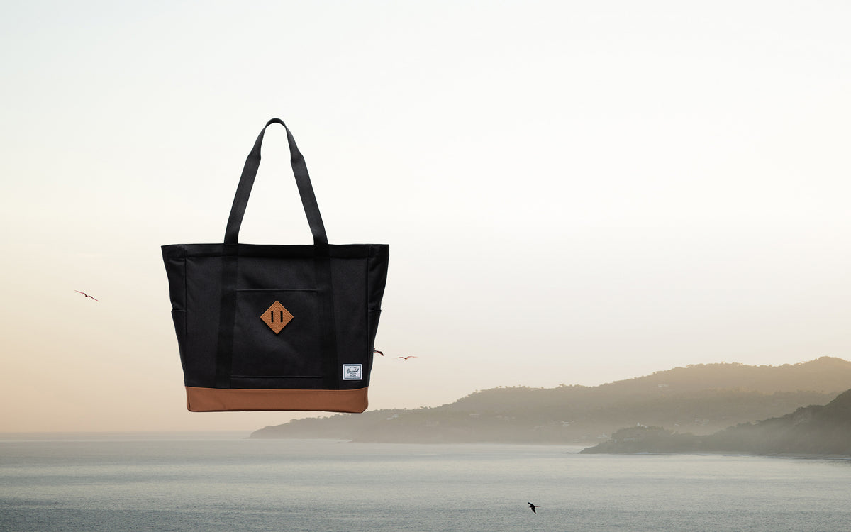 Herschel Heritage Tote bag in Black with Saddle Brown straps and base detailing, displayed on a desktop-optimized hero banner.