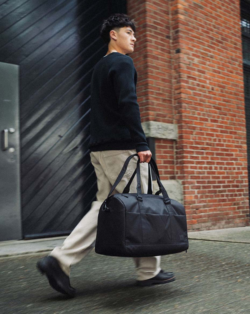 Herschel Supply Co. Novel Duffle Tech in black shown from a side view on a clean background to highlight its durable design and tech organization.