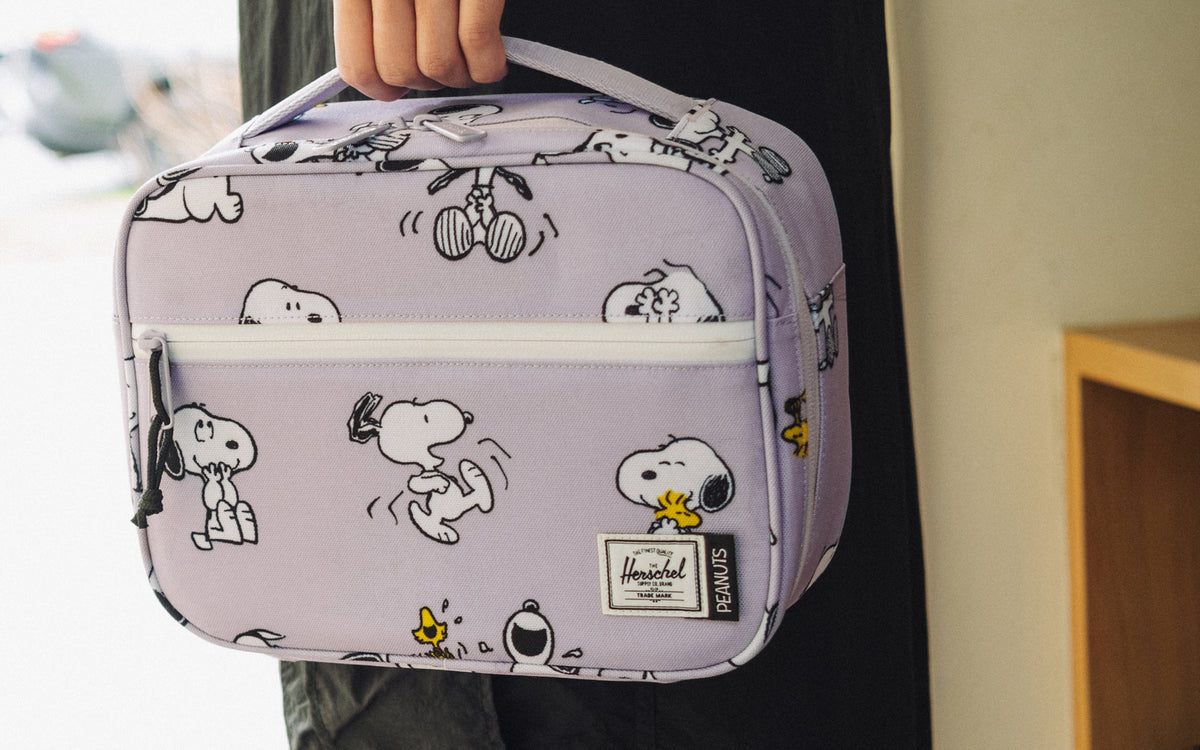 Herschel Supply Co. Peanuts Pop Quiz Lunch Box in Snoopy and Woodstock Pastel Lilac featuring a colorful graphic on a purple background.
