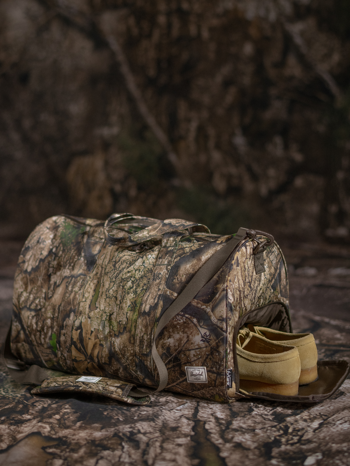 Herschel Supply Co. x Realtree limited-edition collection essential bags, apparel, and accessories arranged together in the APX camouflage pattern.